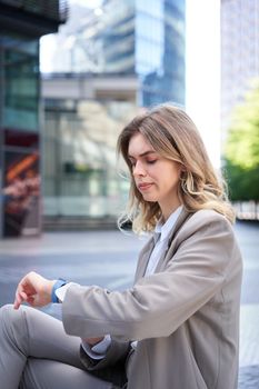 Frowning businesswoman in suit, looking at her digital watch with disappointed face, annoyed waiting for someone, sitting outside
