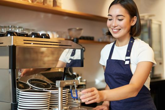 Smiling girl barista in cafe, preparing cappuccino in coffee machine, steaming milk, wearing uniform apron