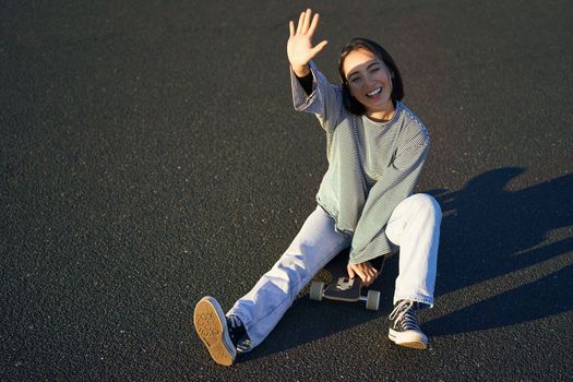 Positive korean girl covers her face from sunlight, sits on skateboard and smiles happily
