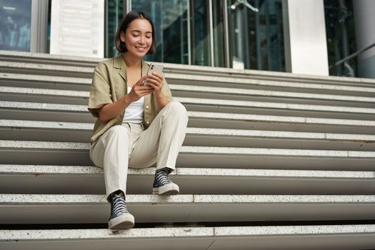 Cellular technology and people. Young happy asian girl sits with smartphone in front of building. Woman using mobile phone, smiling while looking at screen