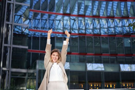 Happy businesswoman dancing on street, raising hands up. Corporate woman celebrates her victory or success