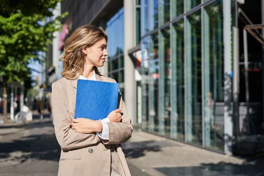 Portrait of businesswoman in beige suit and high heels walking on street. Corporate woman going to work, holding folder with documents