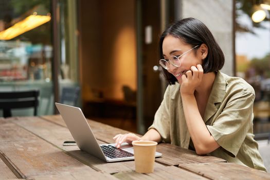Portrait of smiling girl in glasses, sitting with laptop in outdoor cafe, drinking coffee and working remotely, studying online