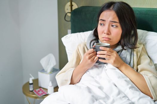 Covid-19, flu and vaccination concept. Korean girl lying in bed with cold, catching influenza, drinking hot tea and using prescribed medication