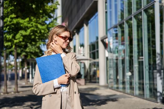 Young saleswoman going to work in sunglasses and suit, holding folder with documents, walking on street on sunny day