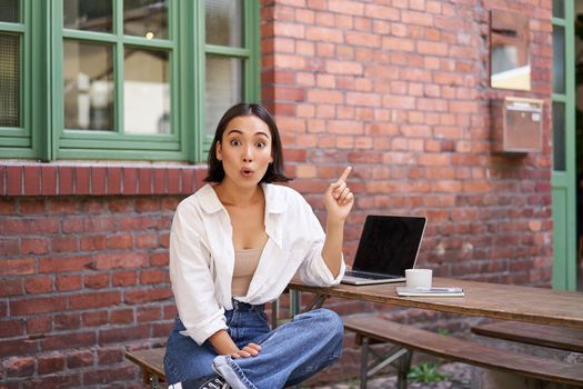 stylish modern asian girl with laptop, sitting in cafe, looking amazed and pointing at upper right corner banner, showing info advertisement