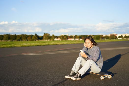 Skater girl sits on her skateboard on road, using smartphone, chatting on mobile app