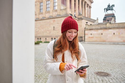 Tourism and sightseeing concept. Young redhead woman, tourist walks around city, looks at her smartphone app and at history stand, explores adventures