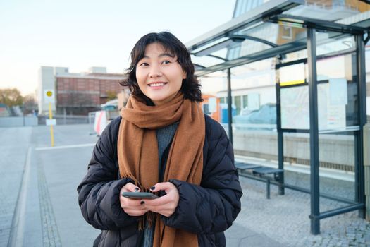 Happy smiling korean girl, using mobile phone, standing on bus stop with smartphone, looking at departure schedule on application, posing in winter clothes