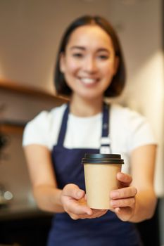 Smiling asian barista girl, giving takeaway coffee cup, prepare takeout order to guest in cafe, wearing apron uniform