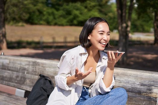 Portrait of happy asian girl records voice message, talking via speakerphone on smartphone, sitting relaxed on bench in park on sunny day