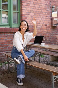 Vertical shot of happy girl attracting attention, waving hand at friend in cafe, sitting on bench with laptop