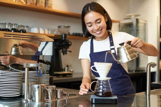 Cute brunette girl barista, cafe staff pouring water from kettle and brewing filter coffee behind counter, preparing order