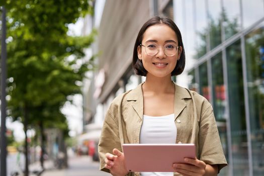 Outdoor shot of young korean woman stands on street with laptop, wears glasses, reads, smiles happily
