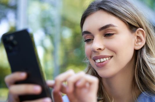 Smiling girl sitting outdoors, looking at smartphone, order on mobile phonr, using application