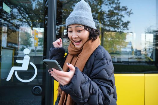 Modern people and lifestyle. Happy asian girl screams from joy, celebrates, stands near bus public transport and looks amazed.