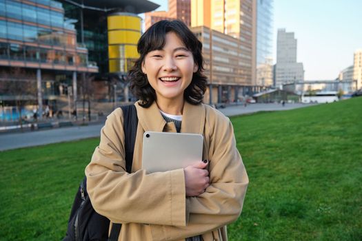 Image of korean girl with happy face, walks around town with student tablet, stands on street and smiles