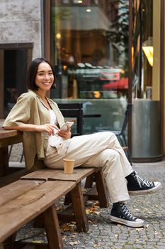 Portrait of stylish young korean woman sits in cafe, holds smartphone, smiles, enjoys coffee outdoors