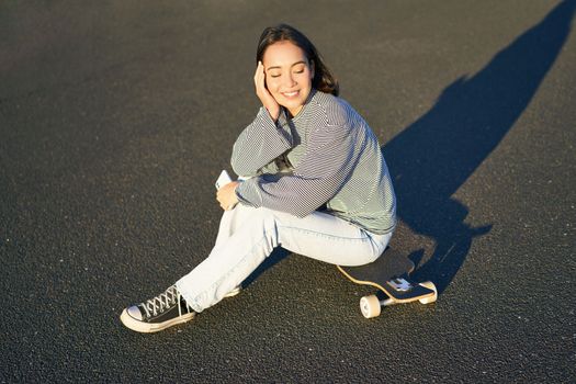 Portrait of asian woman sitting on skateboard, skating on her cruiser longboard, using smartphone app