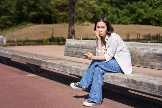 Depressed asian girl sits on bench in park with smartphone, feeling uneasy and stressed, frowning and sighing