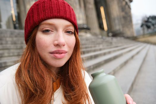 Young redhead female tourist rests during her trip, opens thermos and drinks hot tea, having a break after sightseeing