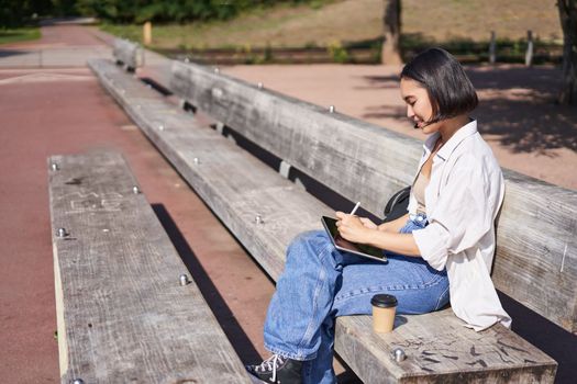 Portrait of asian girl sits on bench in park, draw on her digital tablet with pen, smiling happily, getting creative