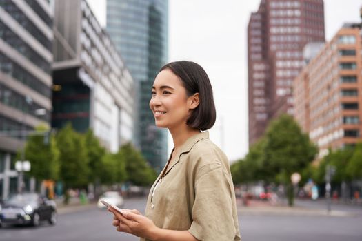 Young asian woman exploring city with smartphone app, holding mobile phone and walking on screet