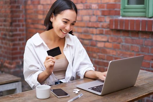 Smiling asian woman sitting with laptop, paying by credit card for online shopping, sending her bank account details, sitting in cafe