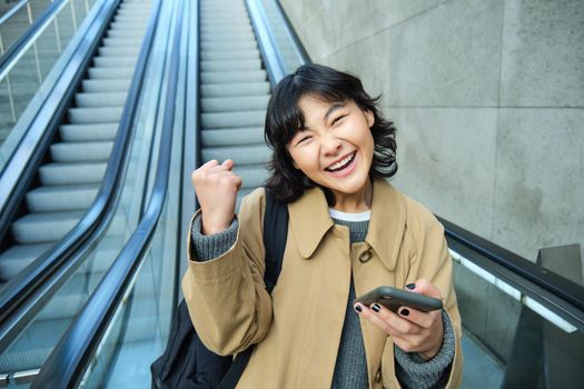 Joyful and positive korean girl, celebrates, looks surprised, goes down escalator with smartphone and looks amazed by smth
