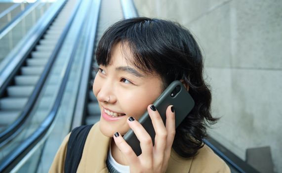 Headshot of smiling korean woman with smartphone, makes a phone call, goes down escalator in city, commutes to university