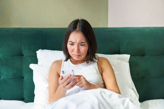 Bedroom picture of asian woman lying in bed, looking scared and shocked at smartphone screen, reading message with concerned face