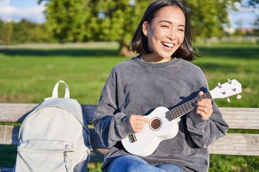 Happy cute girl sits alone on bench in park, plays ukulele guitar and enjoys sunny day outdoors