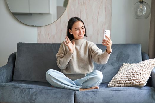 Portrait of asian woman sitting on sofa with smartphone, waving at mobile phone screen and waving at camera, video chat, talking to someone
