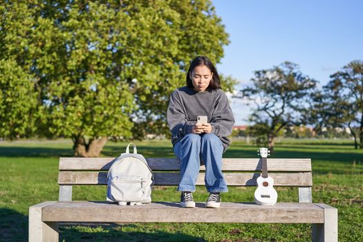 Beautiful brunette girl on bench in park, sitting with ukulele and backpack, holding smartphone, using mobile app