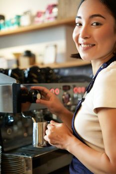 Vertical shot of barista, asian girl steaming milk for cappuccino, prepare latte for client, wearing blue apron, smiling happily, working in cafe