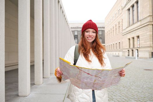 Beautiful redhead woman, tourist with city map, explores sightseeing historical landmark, walking around old town, smiling happily