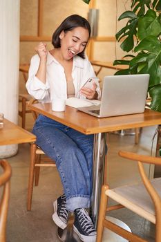 Full body shot of young asian girl sits in cafe with laptop, sees good news on smartphone and celebrates, triumphs from goal or achievement