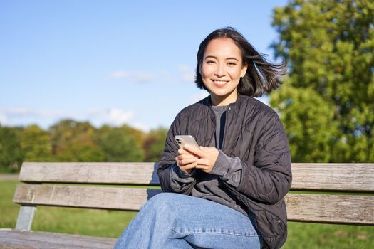 Technology and people. Young woman using smartphone, sitting outdoors on bench in park, looking at smartphone, sending message or networking.