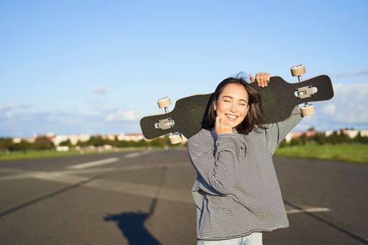 Leisure and people. Happy asian woman standing with longboard, cruising on an empty road in countryside. Skater girl holds her skateboard and smiles at camera