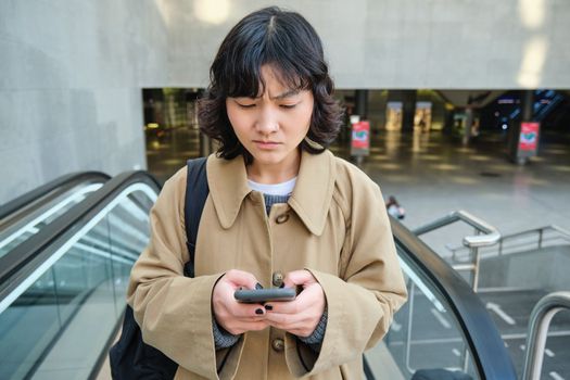 People in city. Portrait of girl looks concerned at smartphone screen with frowned worried face expression. Woman goes up escalator with mobile phone