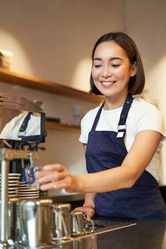 Portrait of cute barista girl working behind counter, making coffee, steaming milk for cappuccino, wearing cafe uniform blue apron
