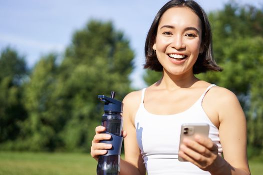 Sportswoman workout with smartphone, drinks water, holds mobile phone and smiles at camera, stands in park