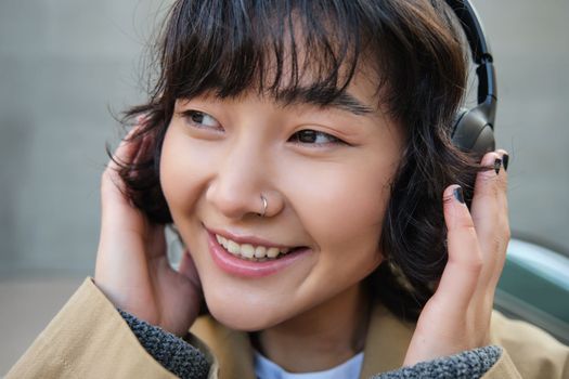 Beautiful smiling korean girl in headphones, wondering around town, standing on street and smiling, listening to music