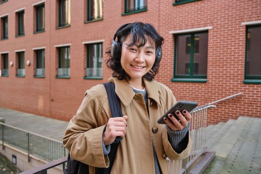 Young happy woman in trench coat, listens music in headphones, holds smartphone, uses mobile application to find route in new city, stands on street with backpack