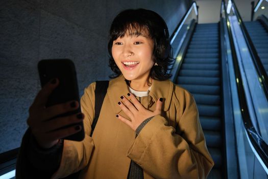 Surprised asian girl looks at smartphone screen, feeling amazed by smth she read on mobile phone, going down escalator in city, commuting to university