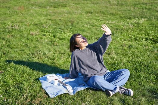 Happy asian girl sitting on picnic blanket with ukulele, cover herself from sunlight, extending hand towards sun beams and smiling