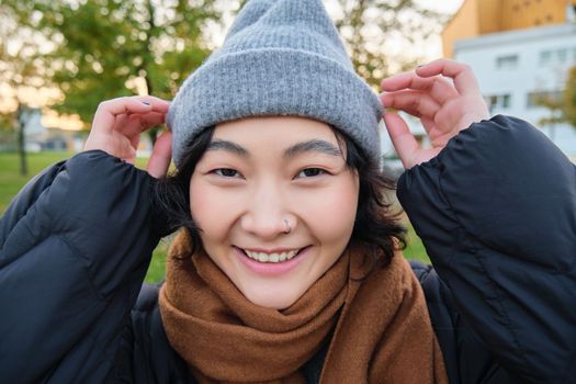 Portrait of cute asian girl in hat and scarf, walks around town in chilly spring weather, smiles and looks happy, sits in park near green grass