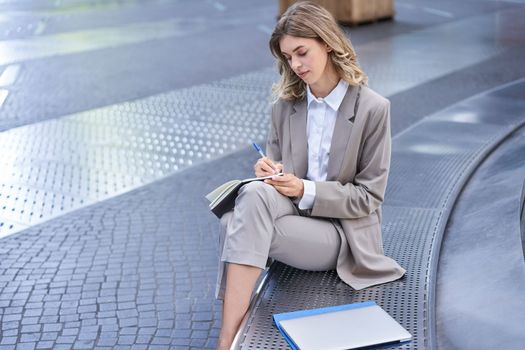 Business woman sits in city centre and works on mobile phone, she has her laptop and wears working corporate outfit