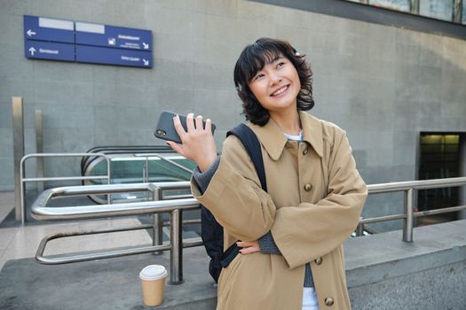 Happy young woman in headphones, stands on street with smartphone and drinks coffee, waits for commute train or bus, travels in city