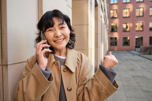 Portrait of smiling happy korean girl, stands on street, receives good news over phone, talks on smartphone and makes fist pump, celebrates, feels excitement
